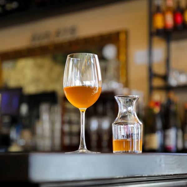 A wine glass and a small carafe, both containing amber liquid, sit on a bar.