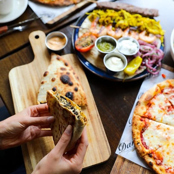 Hands holding a folded flatbread, with a pizza and a Mediterranean platter on a wooden table.