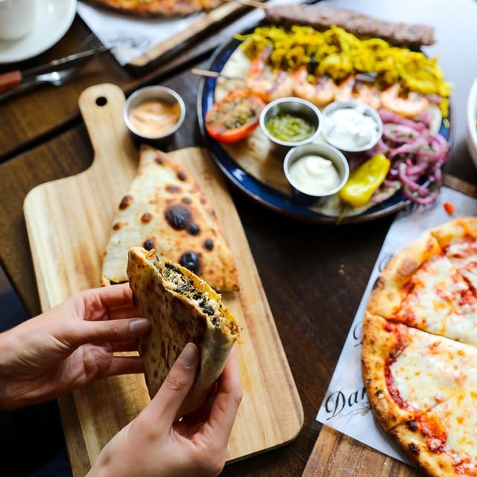 Hands holding a folded flatbread, with a pizza and a Mediterranean platter on a wooden table.