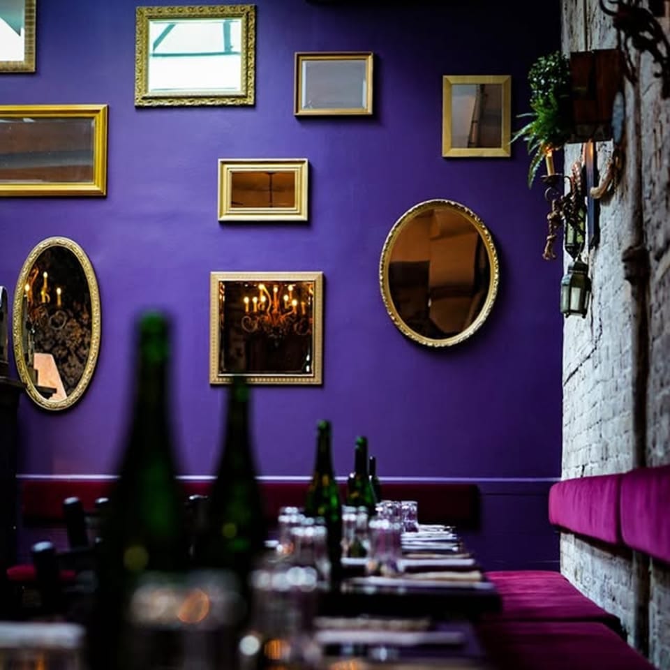 A vibrant purple wall adorned with various gold-framed mirrors, above a dining table with bottles.