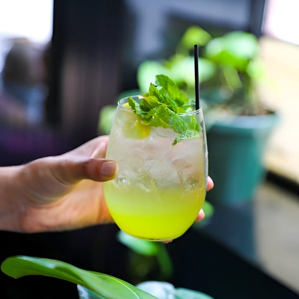 Hand holding a refreshing green cocktail with mint leaves and ice, against a blurred green background.