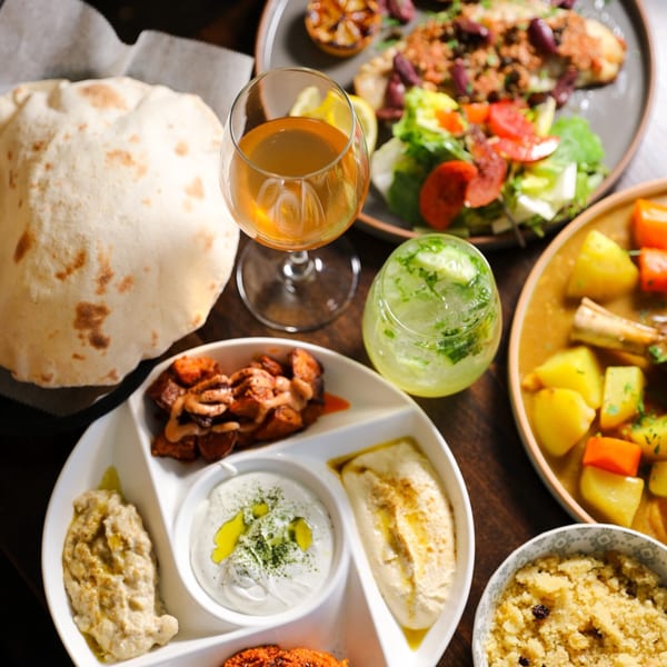 A Mediterranean feast with pita bread, hummus, baba ghanoush, stew, couscous, and drinks.