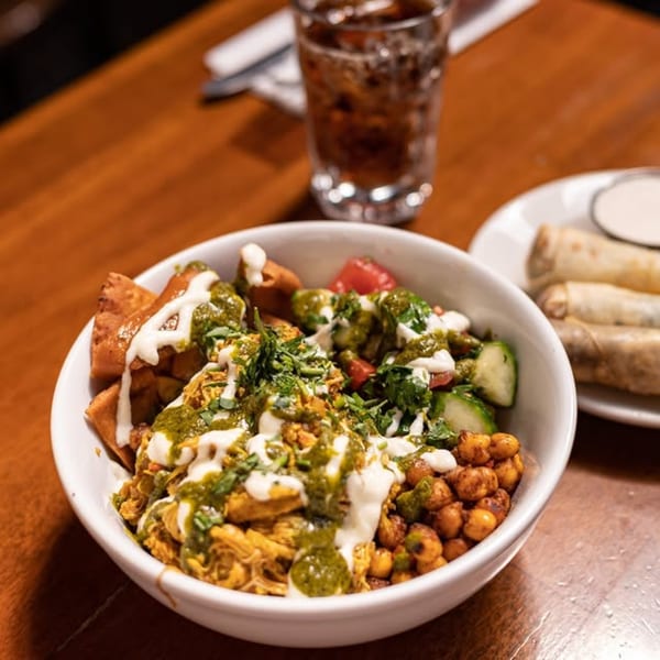 A Mediterranean bowl with chicken, chickpeas, vegetables, and sauces, alongside a drink and spring rolls.