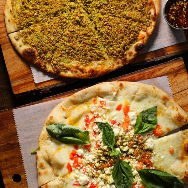 Two Mediterranean pizzas on wooden boards, one with green pistachio topping, the other with cheese, tomatoes, and basil.