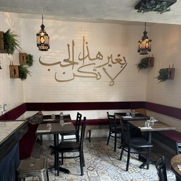 Mediterranean restaurant interior with white brick walls, gold Arabic calligraphy, dark tables, and patterned floor tiles.