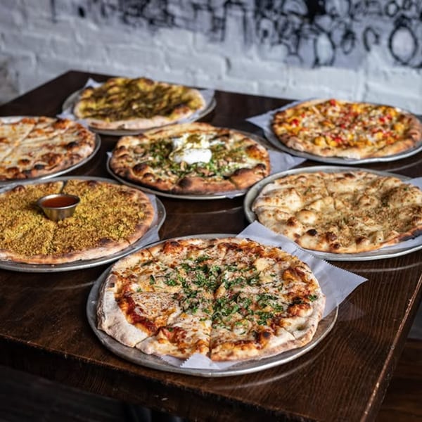 A wooden table laden with seven different Mediterranean flatbreads and pizzas, some with dips.