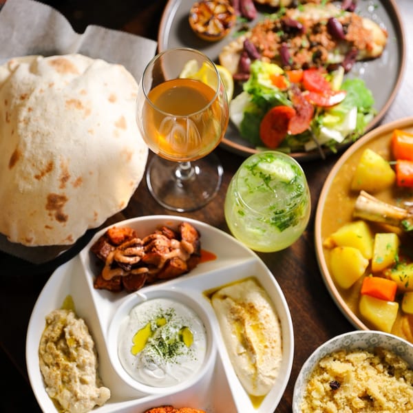 A Mediterranean feast with pita bread, hummus, baba ghanoush, salad, meat, and drinks on a wooden table.