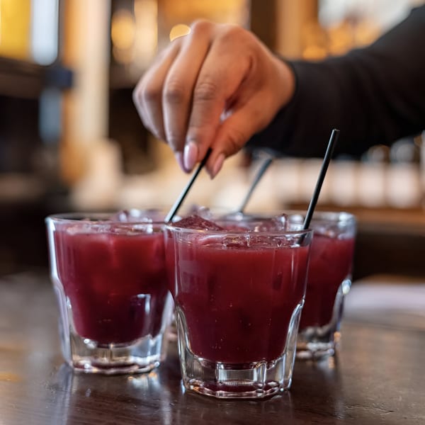 A hand stirs a red cocktail with ice and a black straw on a wooden bar.
