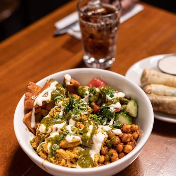 A Mediterranean bowl with chicken, vegetables, chickpeas, and sauces, alongside a drink and spring rolls.
