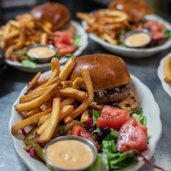 Three plates of Mediterranean burgers with fries, salad, and dipping sauce on a metal counter.
