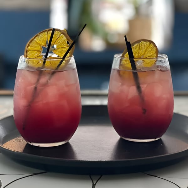 Two vibrant red cocktails with dried orange slices and black straws on a dark tray.