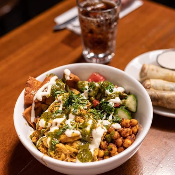 A Mediterranean bowl with chicken, chickpeas, vegetables, and sauces, alongside a drink and spring rolls.