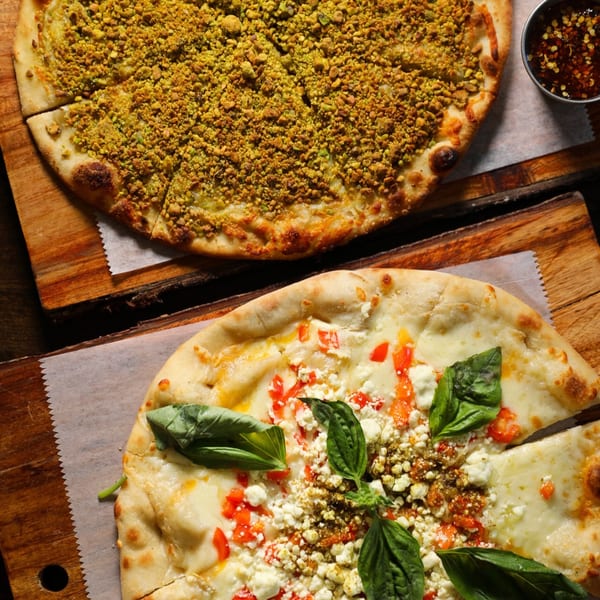 Two Mediterranean pizzas on wooden boards, one with green pistachio topping, the other with cheese, tomatoes, and basil.