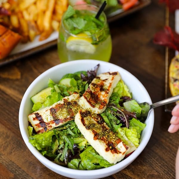 Grilled halloumi cheese salad with mixed greens, cucumber, and a mojito in the background.