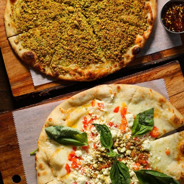 Two Mediterranean pizzas on wooden boards, one with green pistachio topping, the other with cheese, tomatoes, and basil.